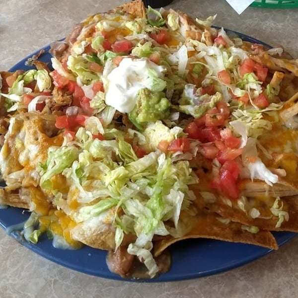 A large plate of Carmela's Mexican Restaurant nachos, topped with cheese, lettuce, tomatoes, guacamole, and sour cream.